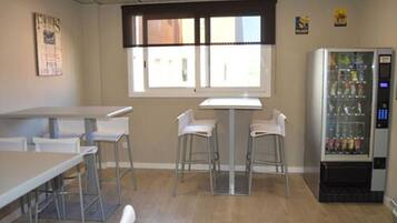 Una habitación con una mesa larga, dos taburetes de bar, una ventana con persianas y una máquina expendedora.