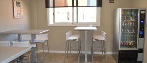 Una habitación con una mesa larga, dos taburetes de bar, una ventana con persianas y una máquina expendedora.