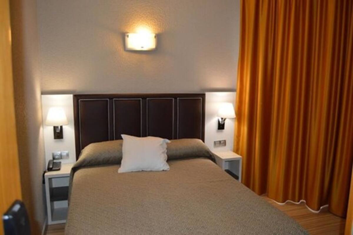 A hotel room with a large bed, two bedside lamps, a headboard, and a curtain.