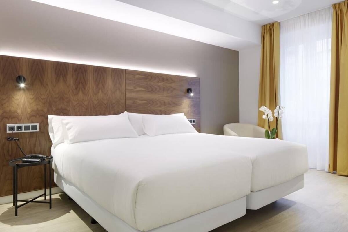 A modern hotel room with a large bed, a bedside table, a chair, and a large window with curtains.