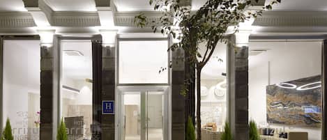 Entrada de hotel con puerta de vidrio, mostrador de recepción y plantas en macetas.