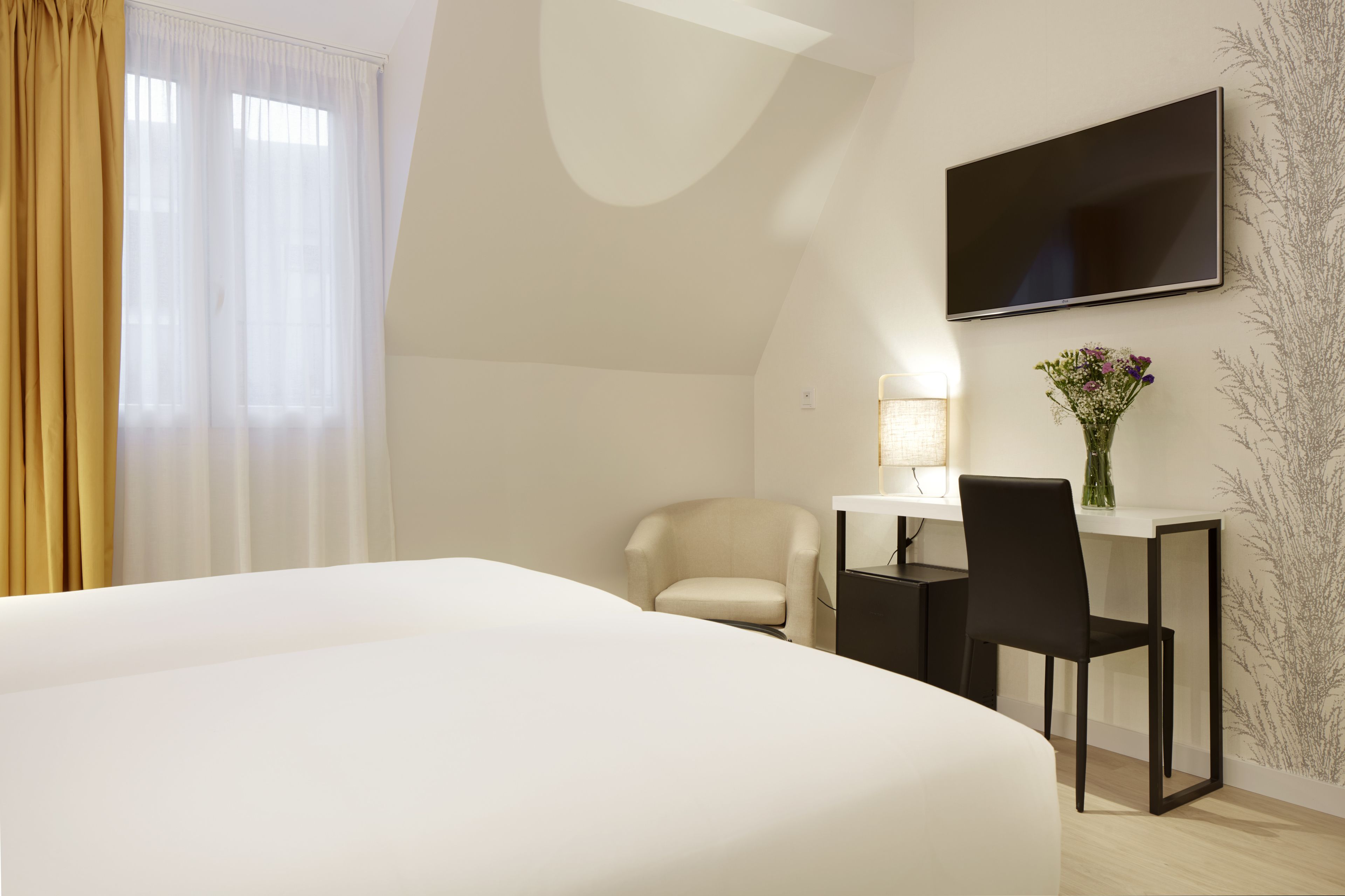 A modern hotel room with a large bed, a desk with a chair, a flat-screen TV, and a vase with flowers.