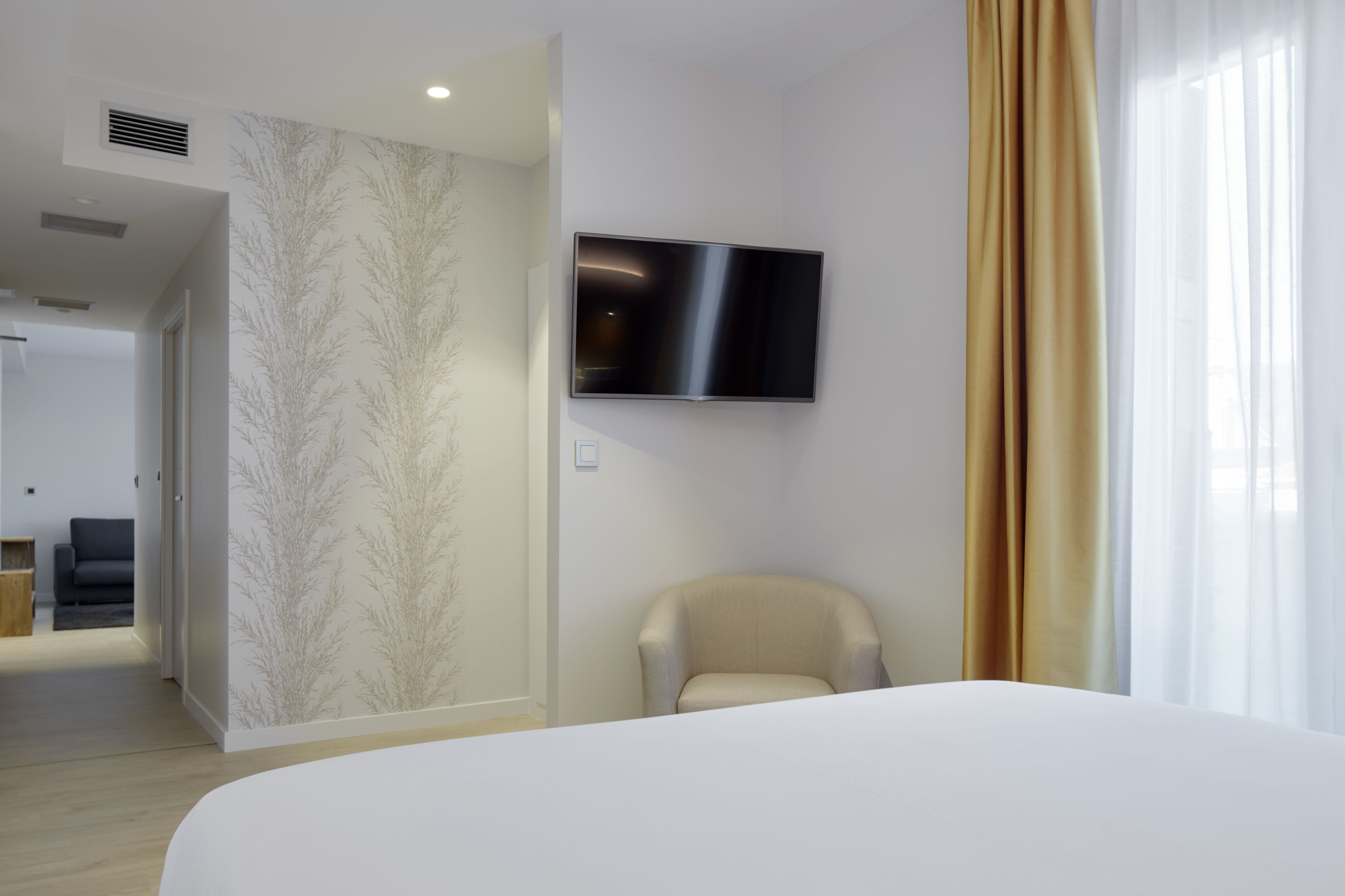 A hotel room with a bed, a TV mounted on the wall, a chair, and a door leading to another room.