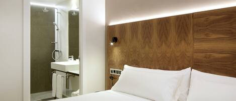 Camera d'albergo moderna con una testiera in legno, un letto bianco e un bagno con doccia e lavandino.