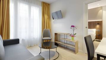 Una habitación de hotel moderna con un sofá, una mesa redonda, un televisor montado en la pared y un pequeño estante de madera con una planta.