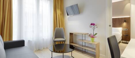 A modern hotel room with a sofa, a round table, a TV mounted on the wall, and a small wooden shelf with a plant.