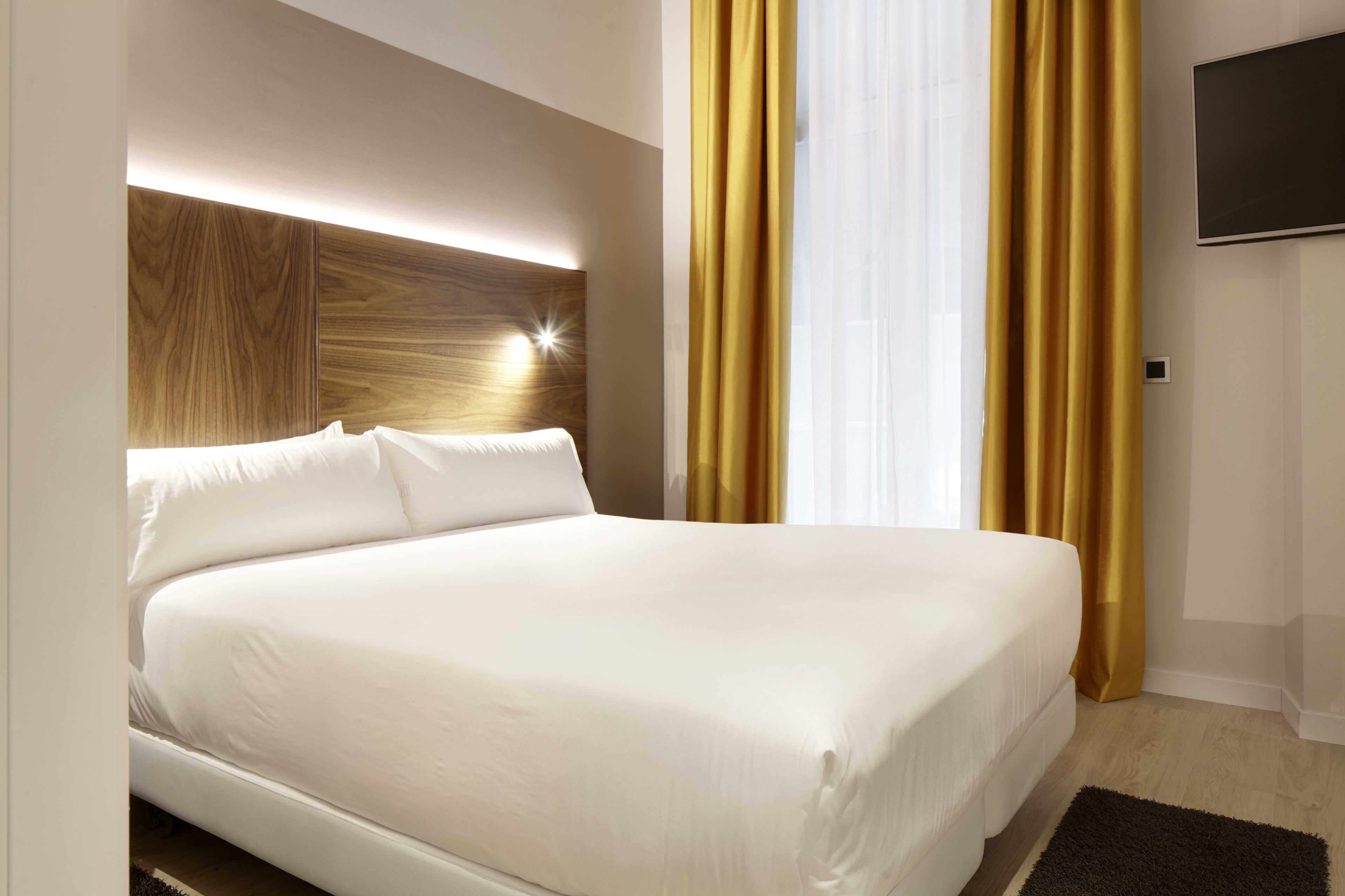 A modern hotel room with a large bed, a wooden headboard, a flat-screen TV, and a large window with yellow curtains.