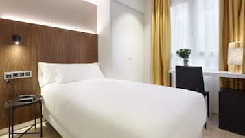 Quarto de hotel com uma cama grande, uma escrivaninha com cadeira, uma mesinha e um abajur.