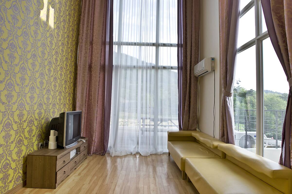 Une pièce comprenant une commode en bois, un téléviseur, un banc et une grande fenêtre avec des rideaux.