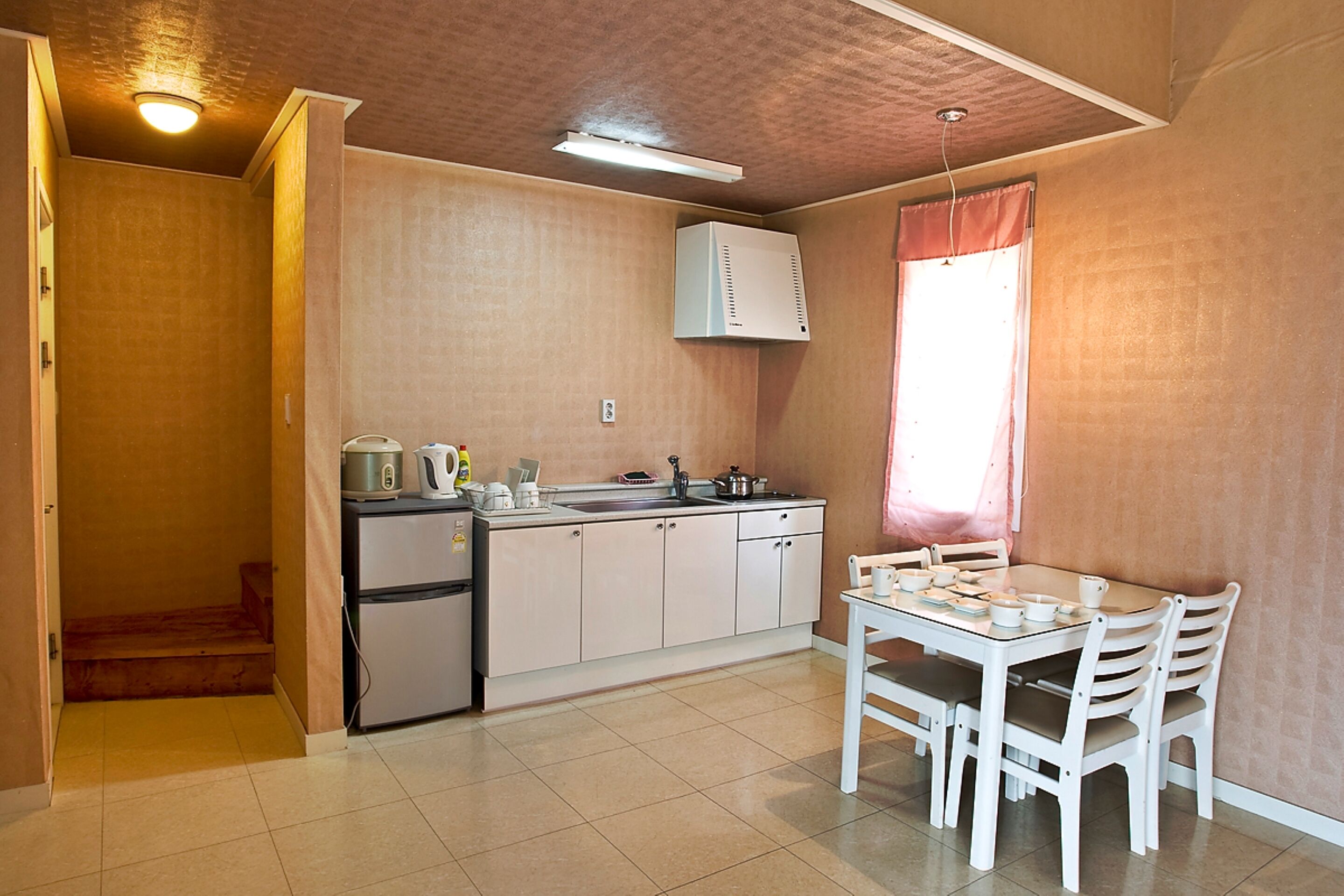 A compact kitchen with a refrigerator, stove, and sink. There is a dining table with chairs and a window with curtains.