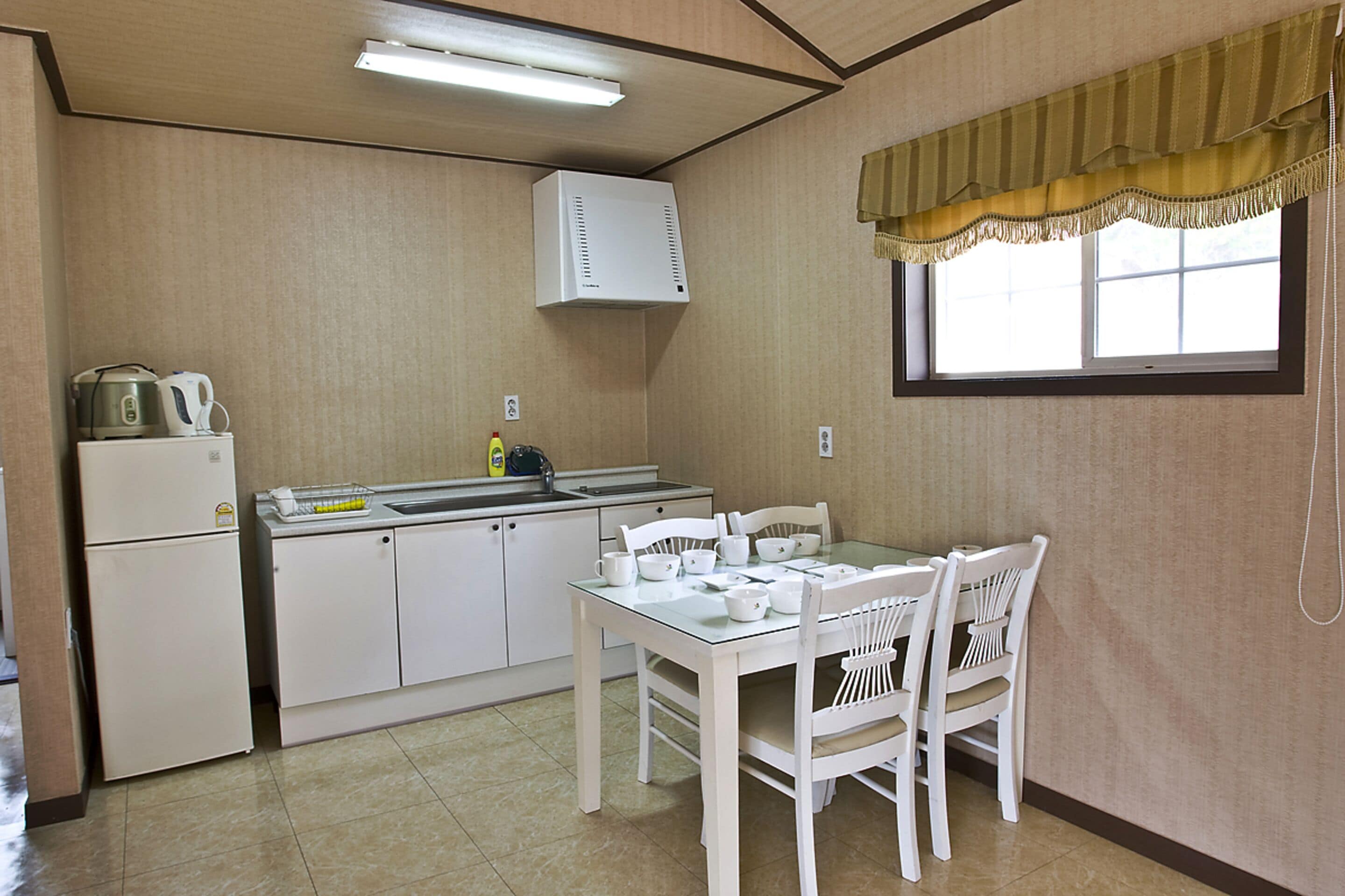 Cocina compacta con refrigerador, estufa y lavamanos. Hay una mesa de comedor con sillas y un ventanal con cortina.