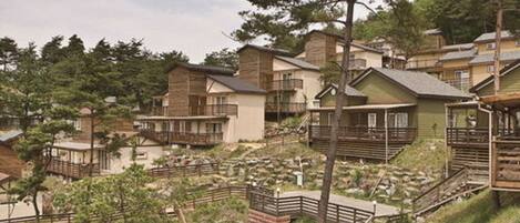 A residential area with houses featuring balconies and wooden railings, surrounded by trees and a hilly landscape.