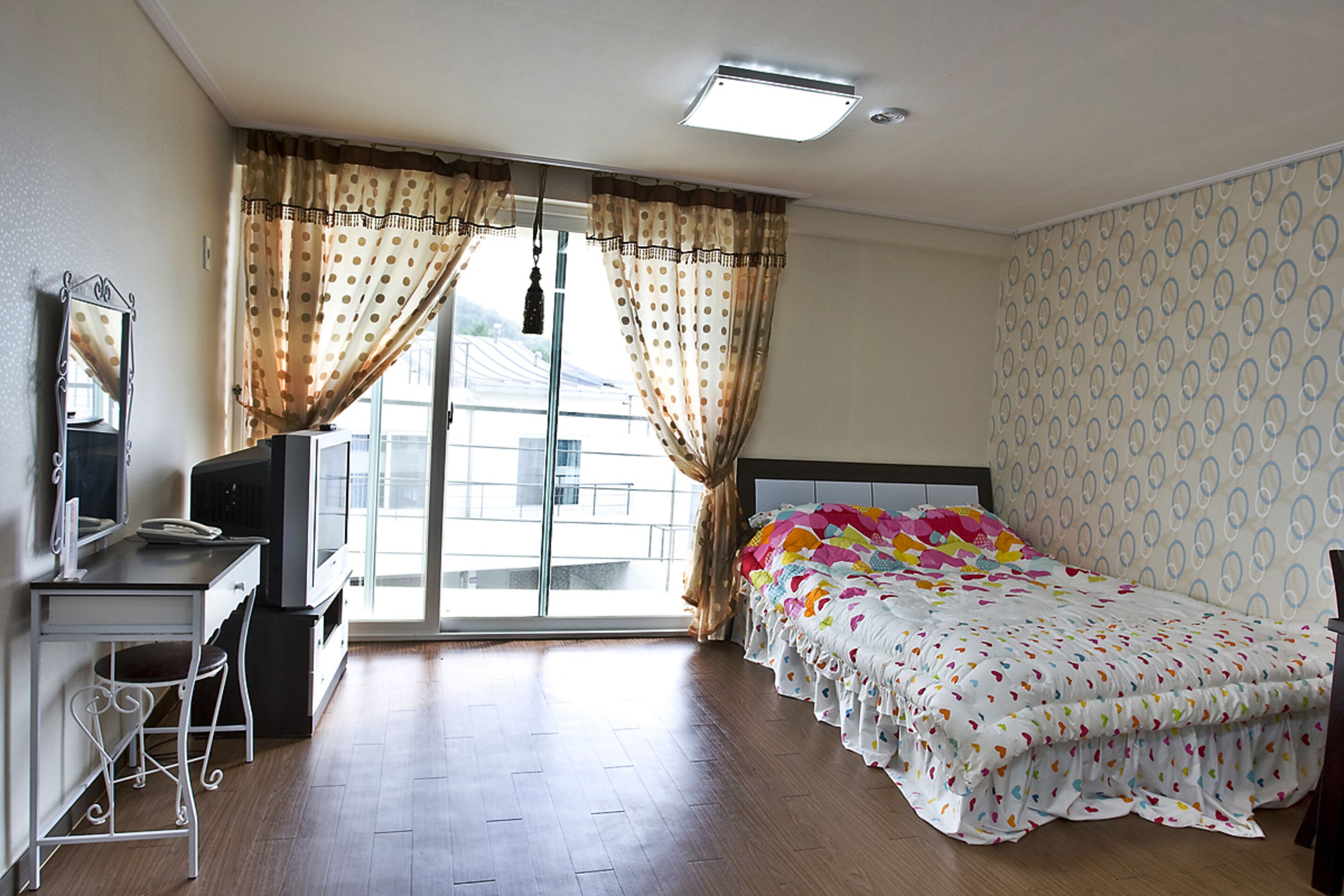 Quarto de hotel com cama, escrivaninha, cadeira, televisão e janela com cortinas.