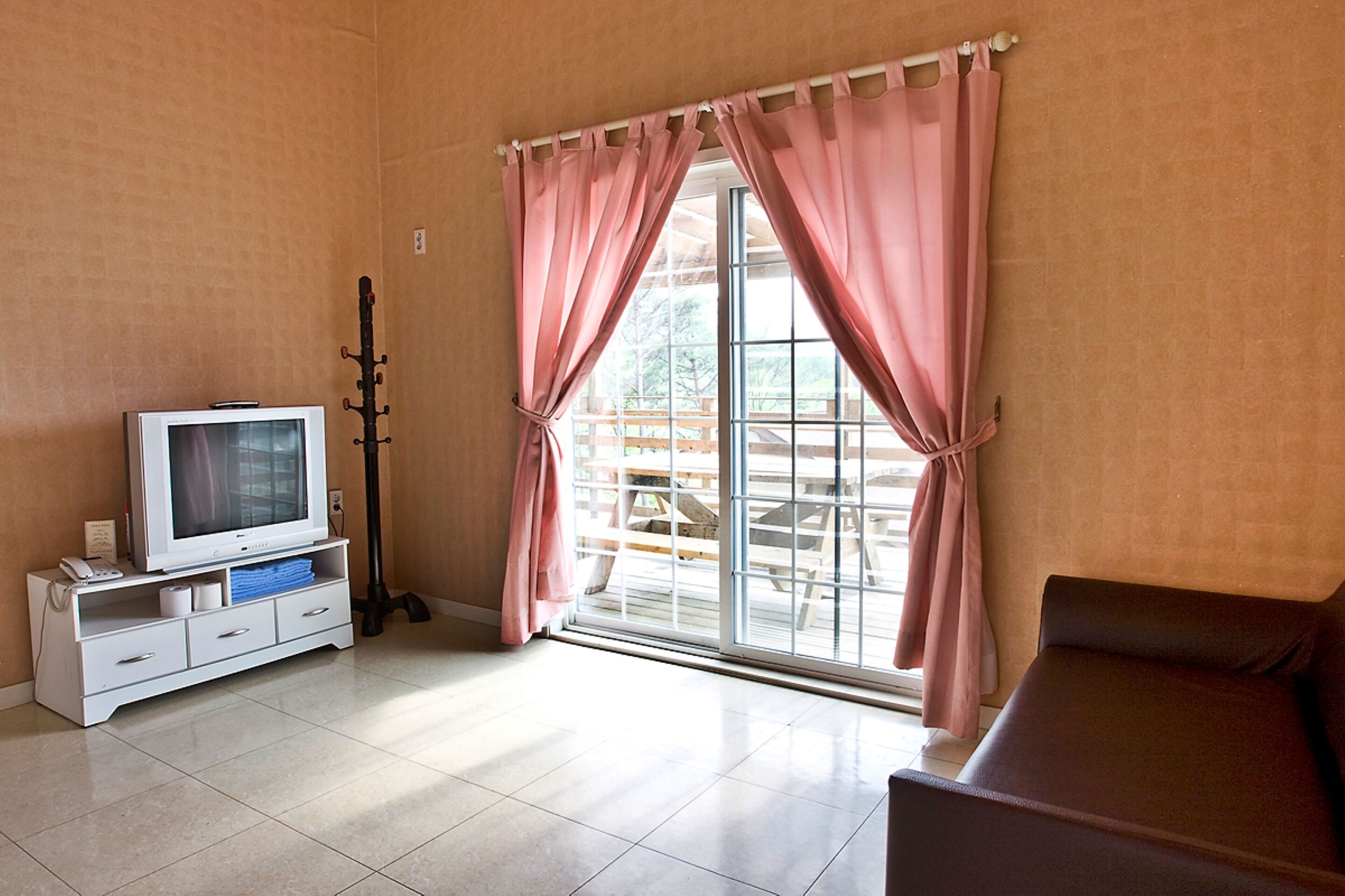 A room with a television, a bench, and a sliding glass door with pink curtains.