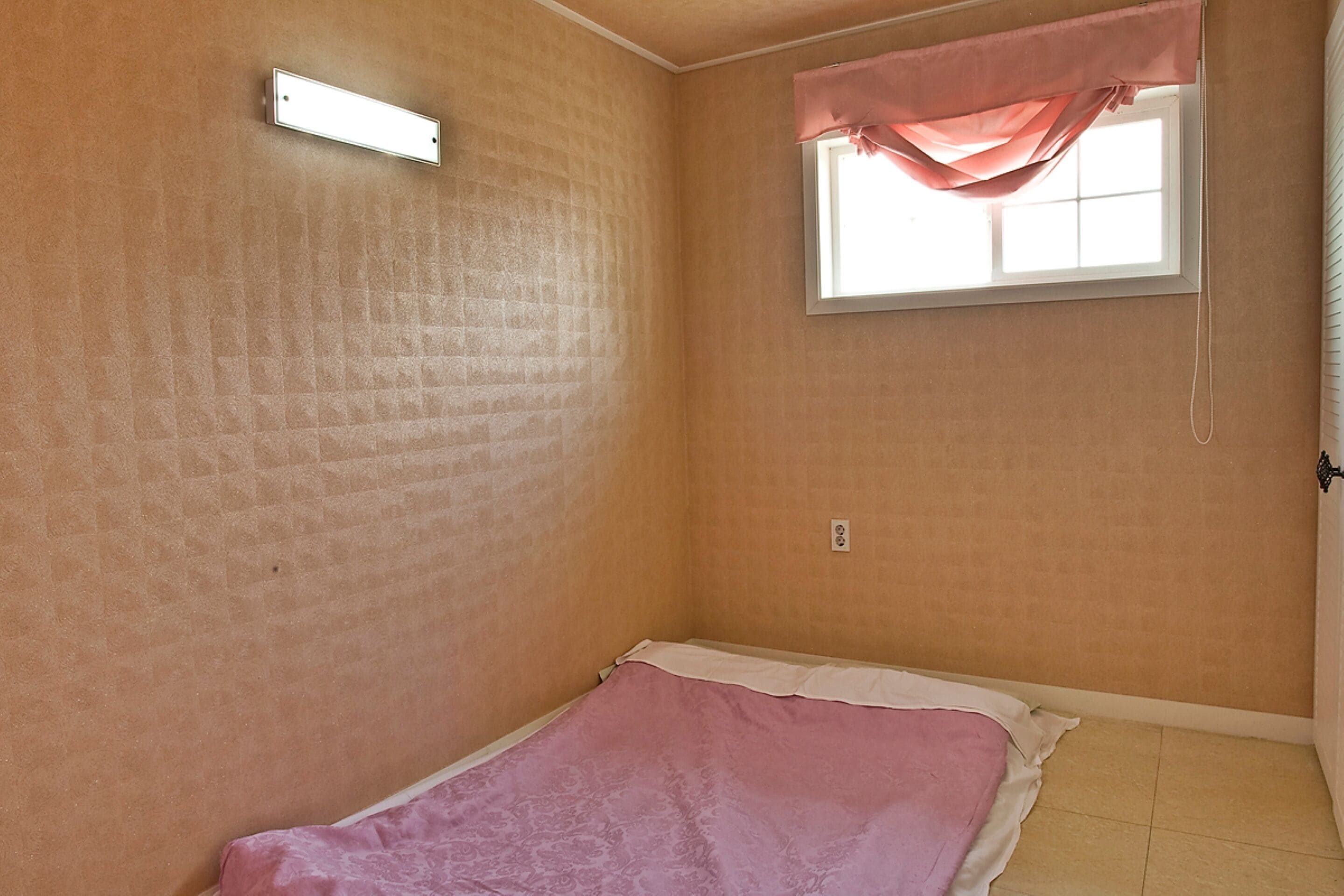 A small, single-room with a bed, a window with a pink curtain, and a wall-mounted light fixture.