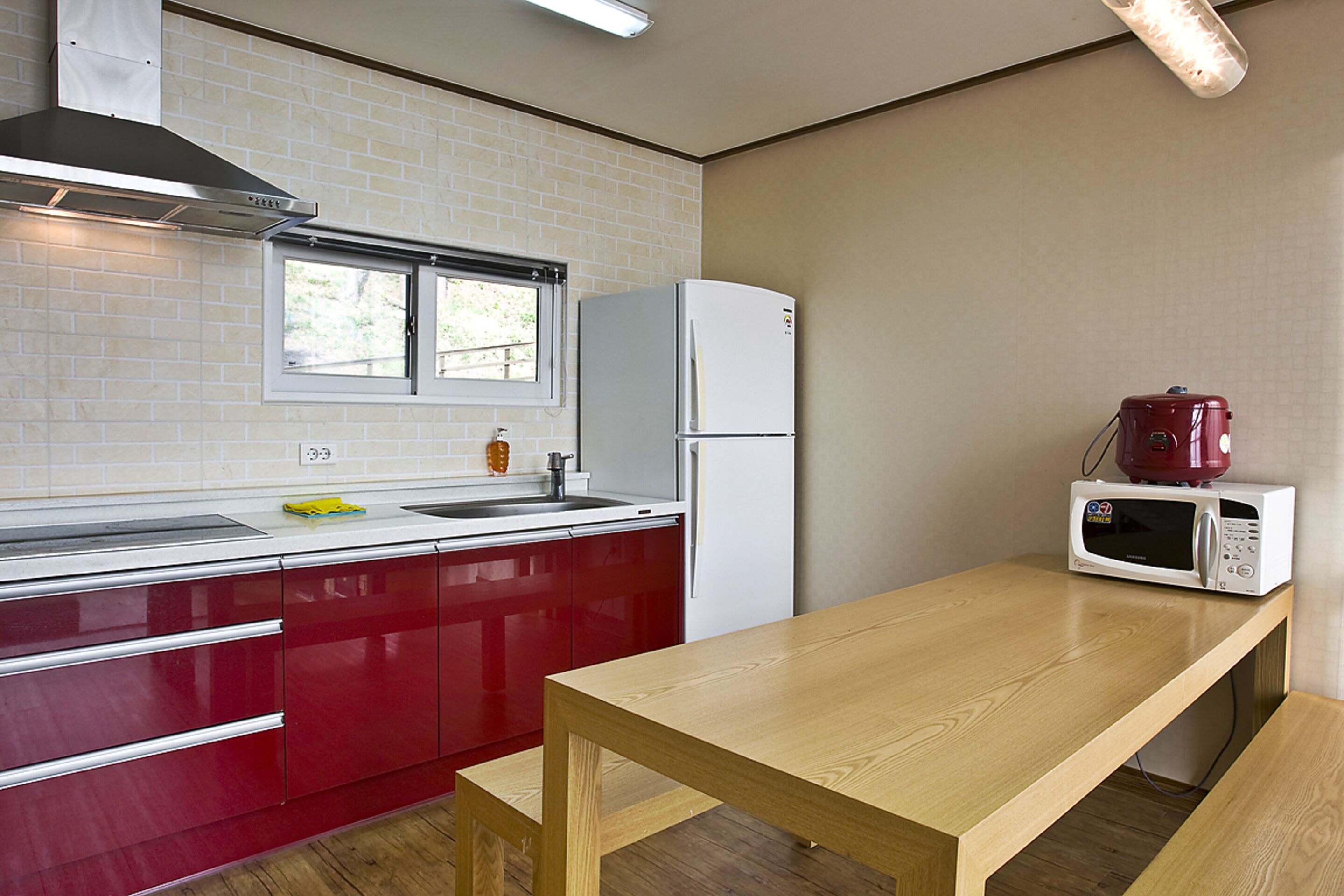 Une cuisine avec des meubles de couleur rouge, un réfrigérateur blanc, une table en bois et un four à micro-ondes posé sur le plan de travail.