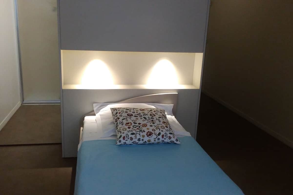 Ein kleines, ordentlich bezogenes Bett mit blauer Bettdecke und einem Blumenkissen, vor einer Wand mit zwei Wandleuchten.
