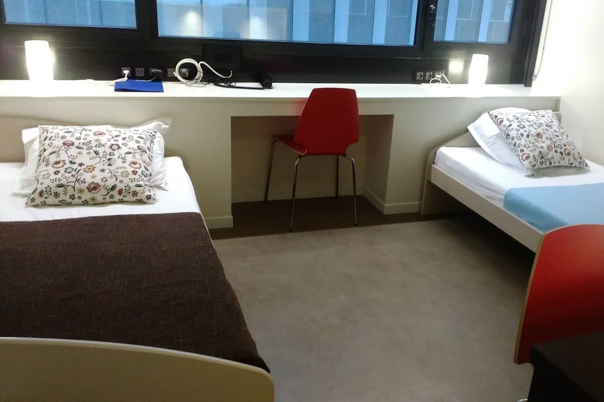 Ein kleiner, kompakter Raum mit zwei Einzelbetten, einem Schreibtisch, einem Stuhl und einem Fenster.