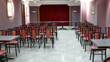 Un salón de conferencias con filas de sillas y mesas, un escenario con cortina roja y obras de arte enmarcadas en las paredes.