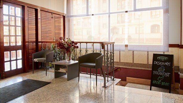 A hotel lobby with a wooden door, a reception desk, a chair, a vase with flowers, and a chalkboard sign advertising breakfast hours. - Hotel San Jorge (Santurtzi)