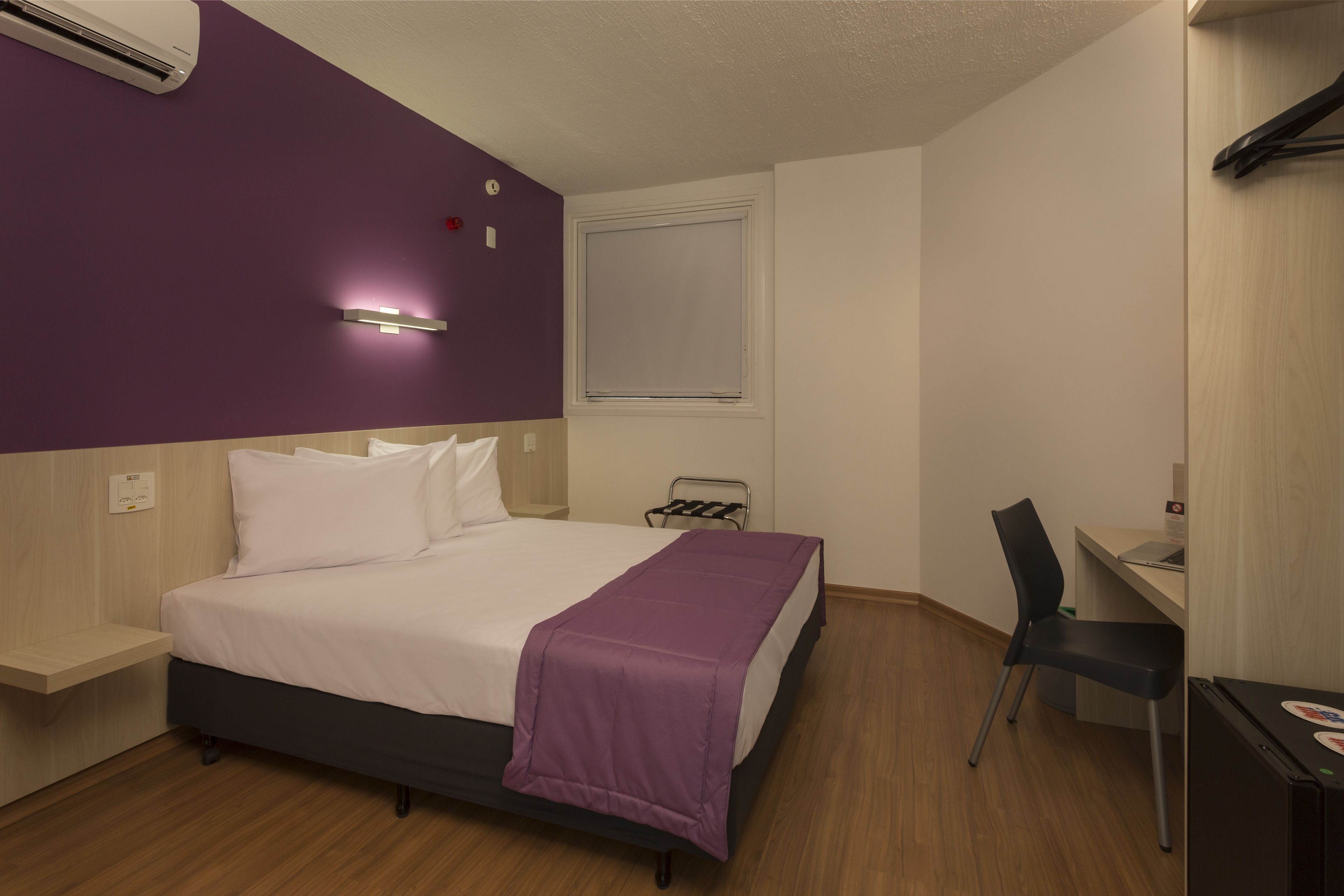 Quarto de hotel com cama, escrivaninha, cadeira e ar condicionado.