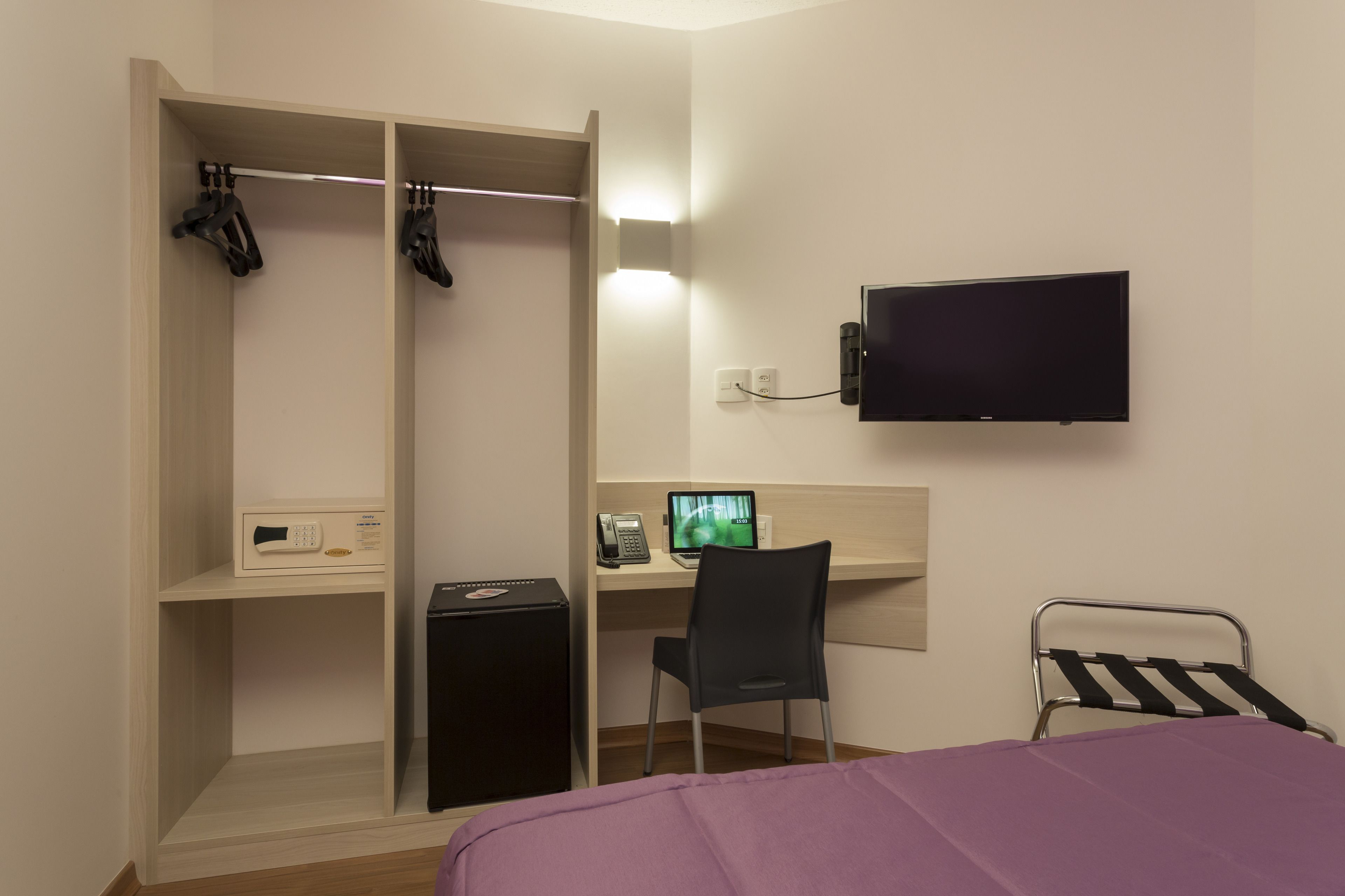 A bedroom with a bed, a desk with a computer, a TV, a wardrobe, and a small fridge.