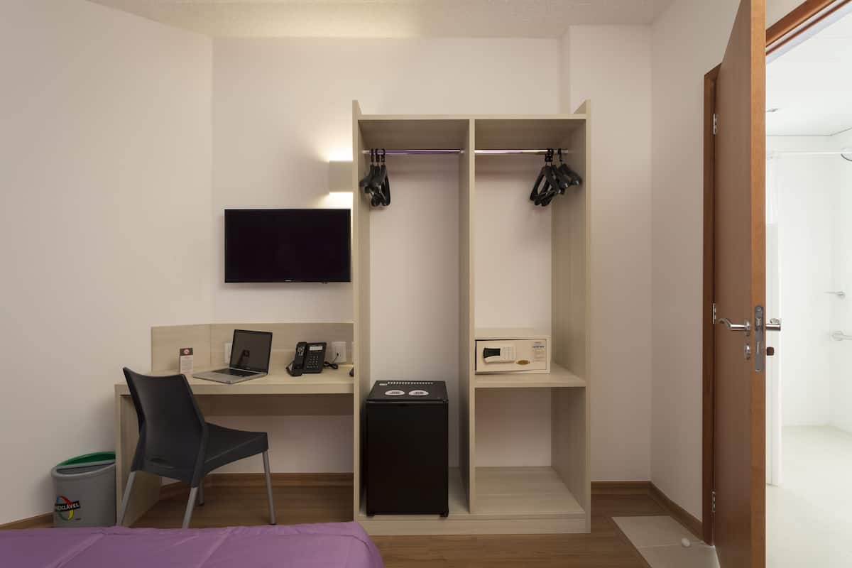 A compact hotel room with a bed, a desk with a laptop, a chair, a TV mounted on the wall, a wardrobe, and a small trash bin.