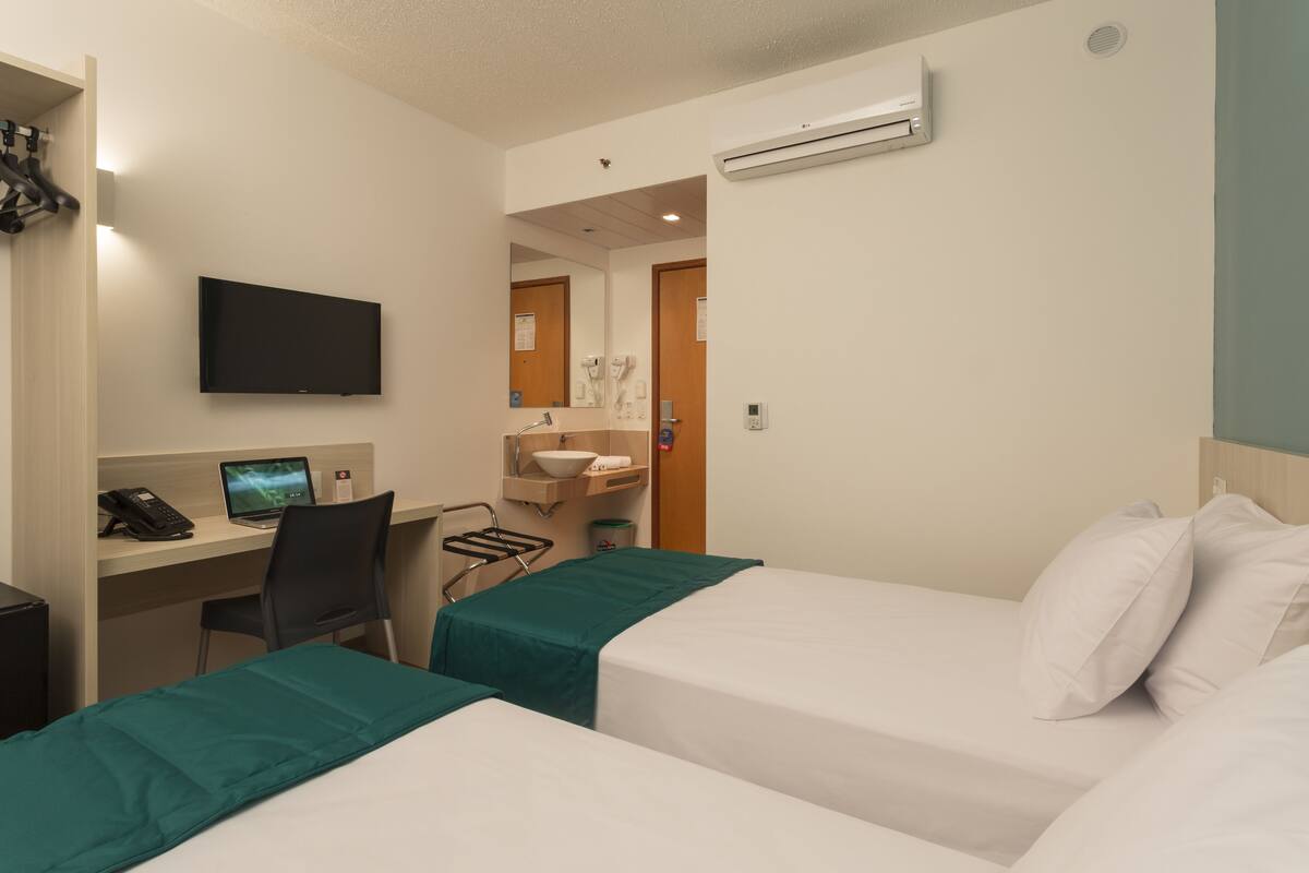 A hotel room with two beds, a desk with a computer, a TV, and a bathroom with a sink and mirror.