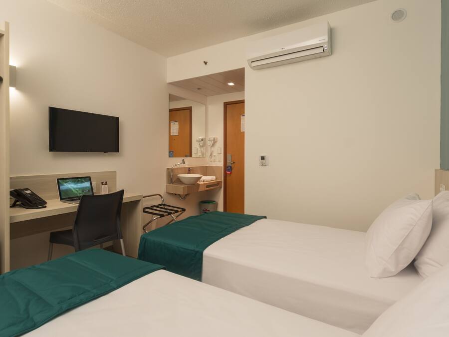 Una habitación de hotel con dos camas, un escritorio con una computadora, un televisor y un baño con lavamanos y espejo.