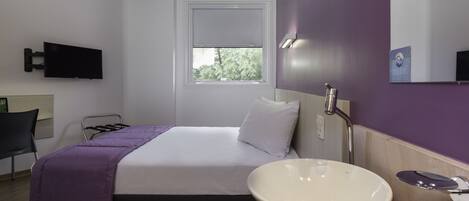 A hotel room with a bed, a bedside table with a white basin, a flat-screen TV mounted on the wall, and a window with a view of trees.
