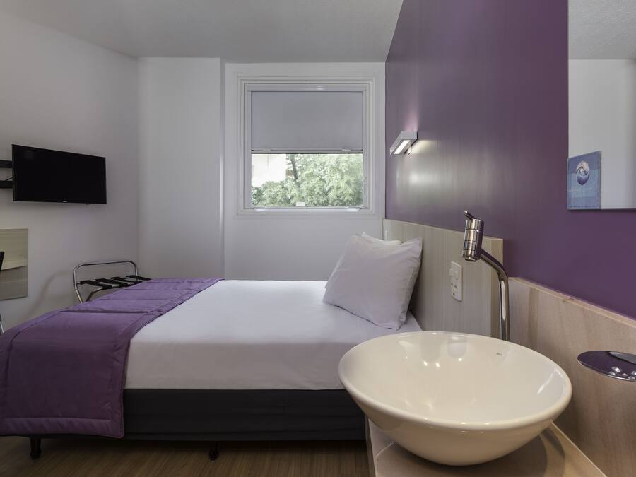 Habitación de hotel con cama, mesita de noche con lavamanos blanco, televisor de pantalla plana en la pared y ventana con vistas a árboles.