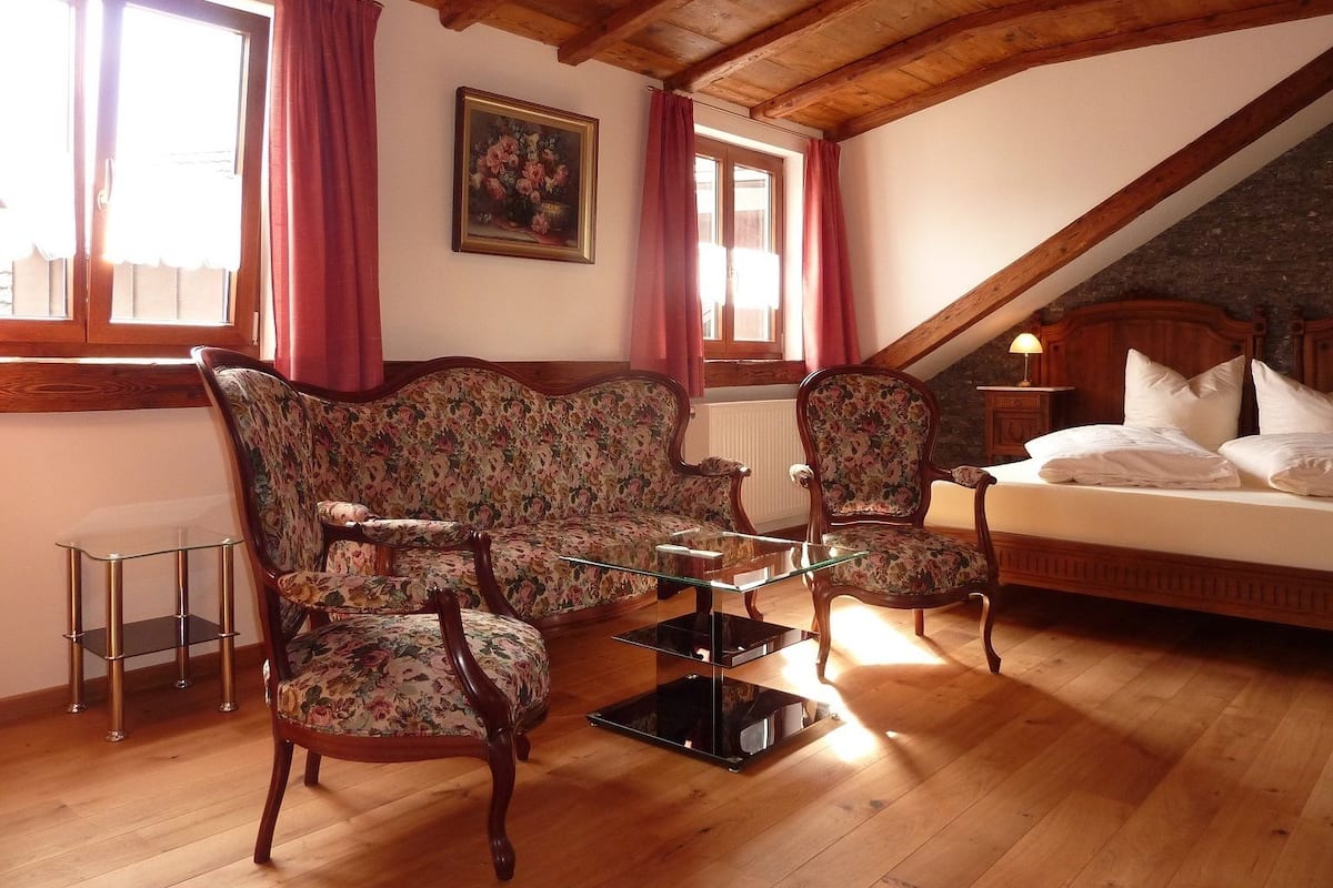 A cozy room with a wooden ceiling, a floral sofa, two armchairs, a glass coffee table, a bed with white linens, and a painting on the wall.