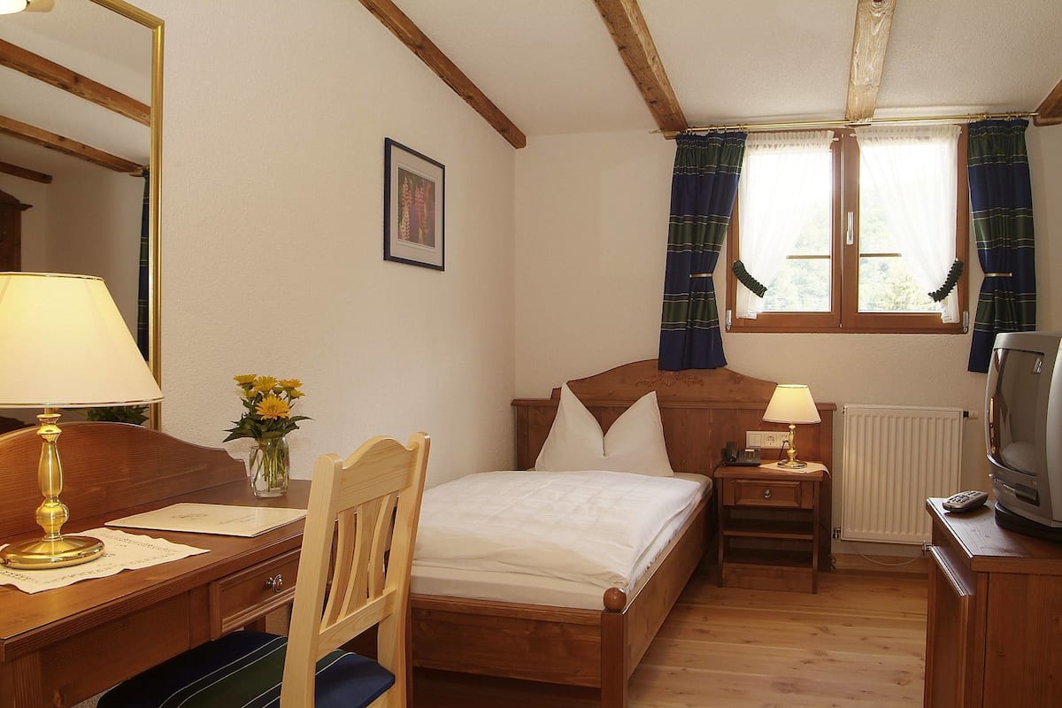 A wooden-framed bedroom with a bed, bedside lamps, a desk, a chair, a television, and a window with curtains.