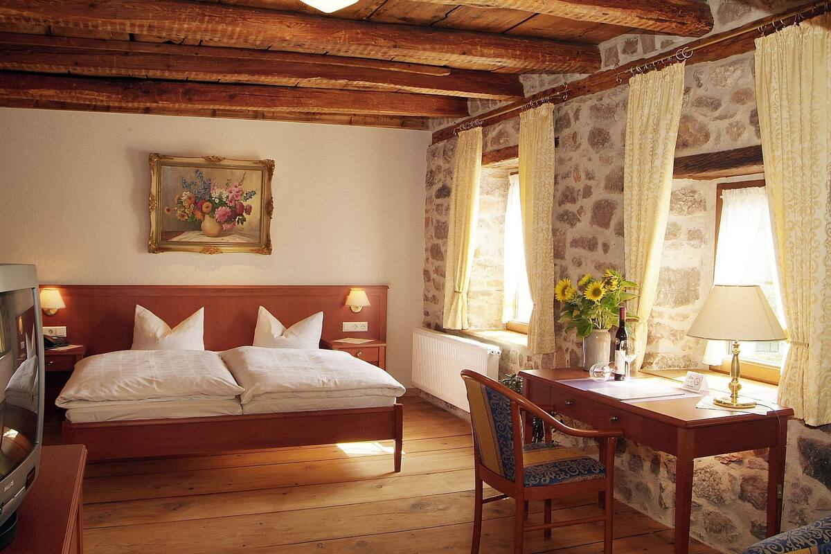 A cozy room with a wooden bed, a desk with a lamp, a chair, and a painting on the wall.