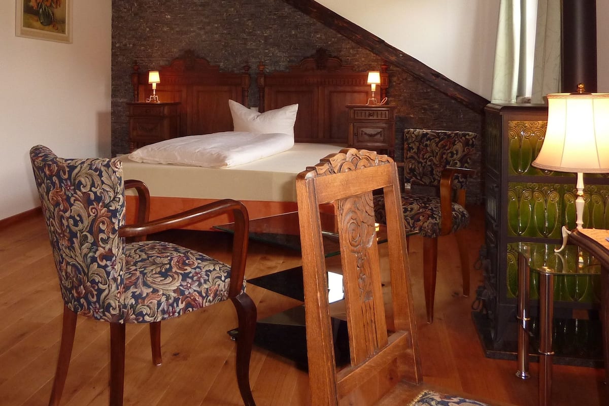 A bedroom with a sloped ceiling, a wooden bed, two upholstered chairs, a nightstand with a lamp, and a green glass table with a lamp.