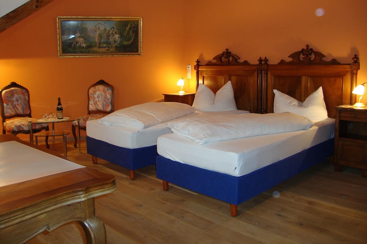 A room with two beds, wooden headboards, a small table with a bottle and glasses, and a painting on the wall.