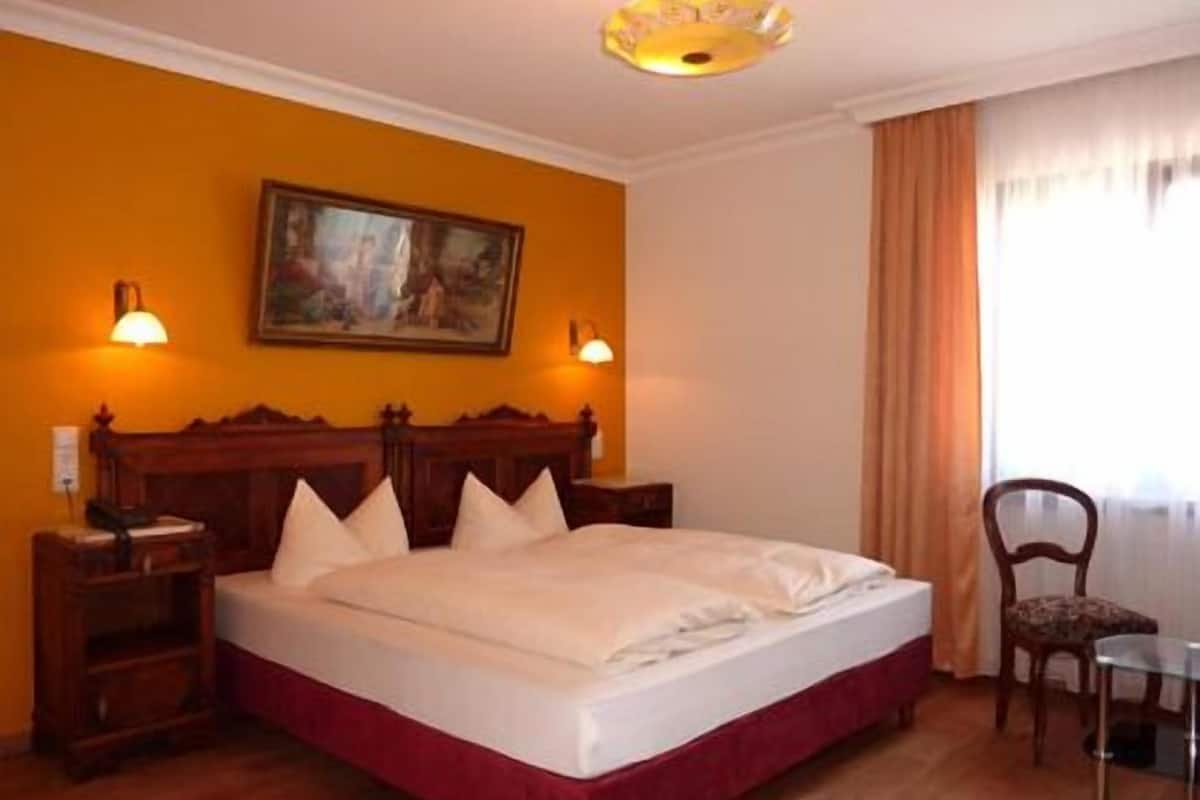 A hotel room with a wooden bed, two bedside tables, a chair, a painting on the wall, and a window with curtains.