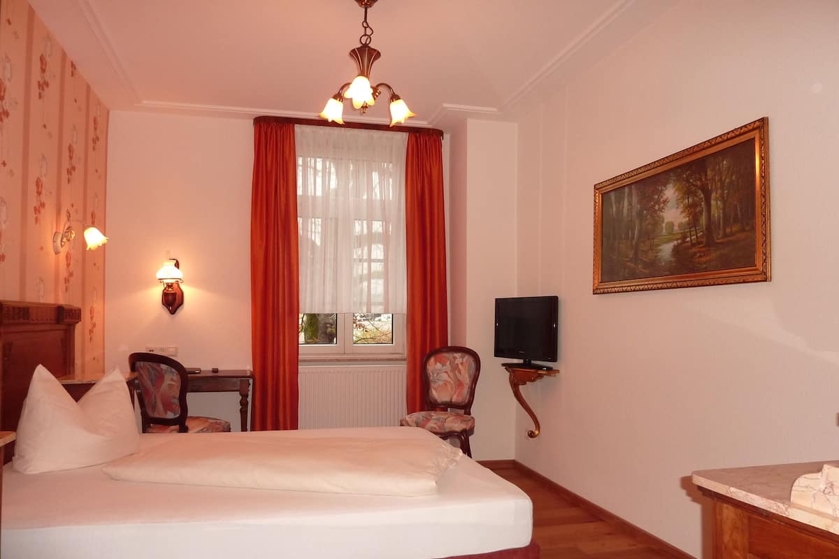 A hotel room with a bed, a desk with a television, a painting on the wall, and a window with curtains.
