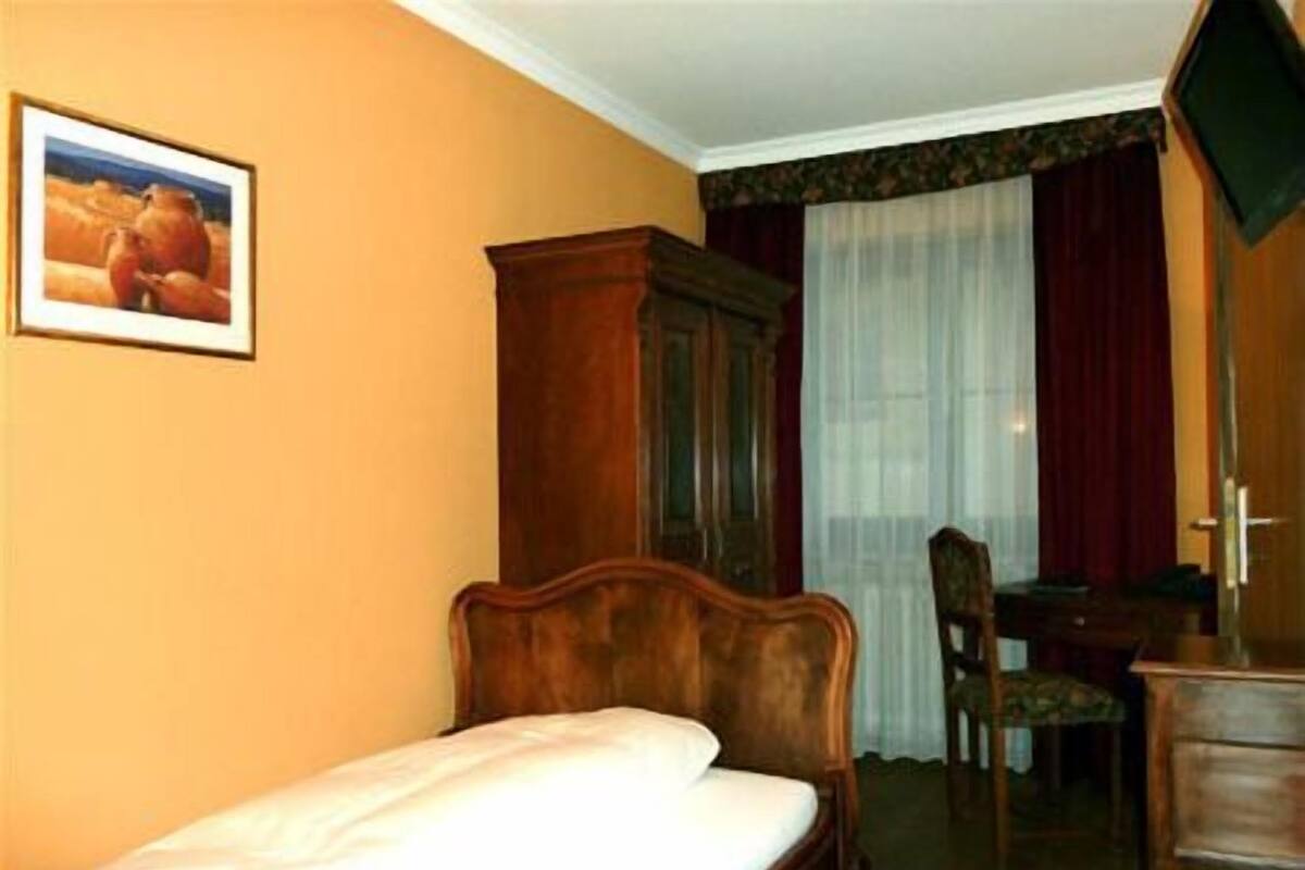 A hotel room with a single bed, a wooden wardrobe, a desk with a chair, and a television.