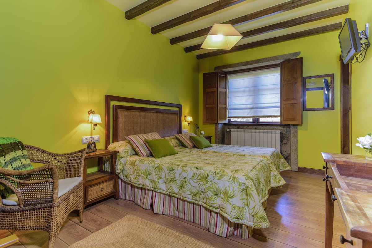 A bedroom with a bed, a wicker chair, a nightstand, a lamp, a window with shutters, and a television mounted on the wall.