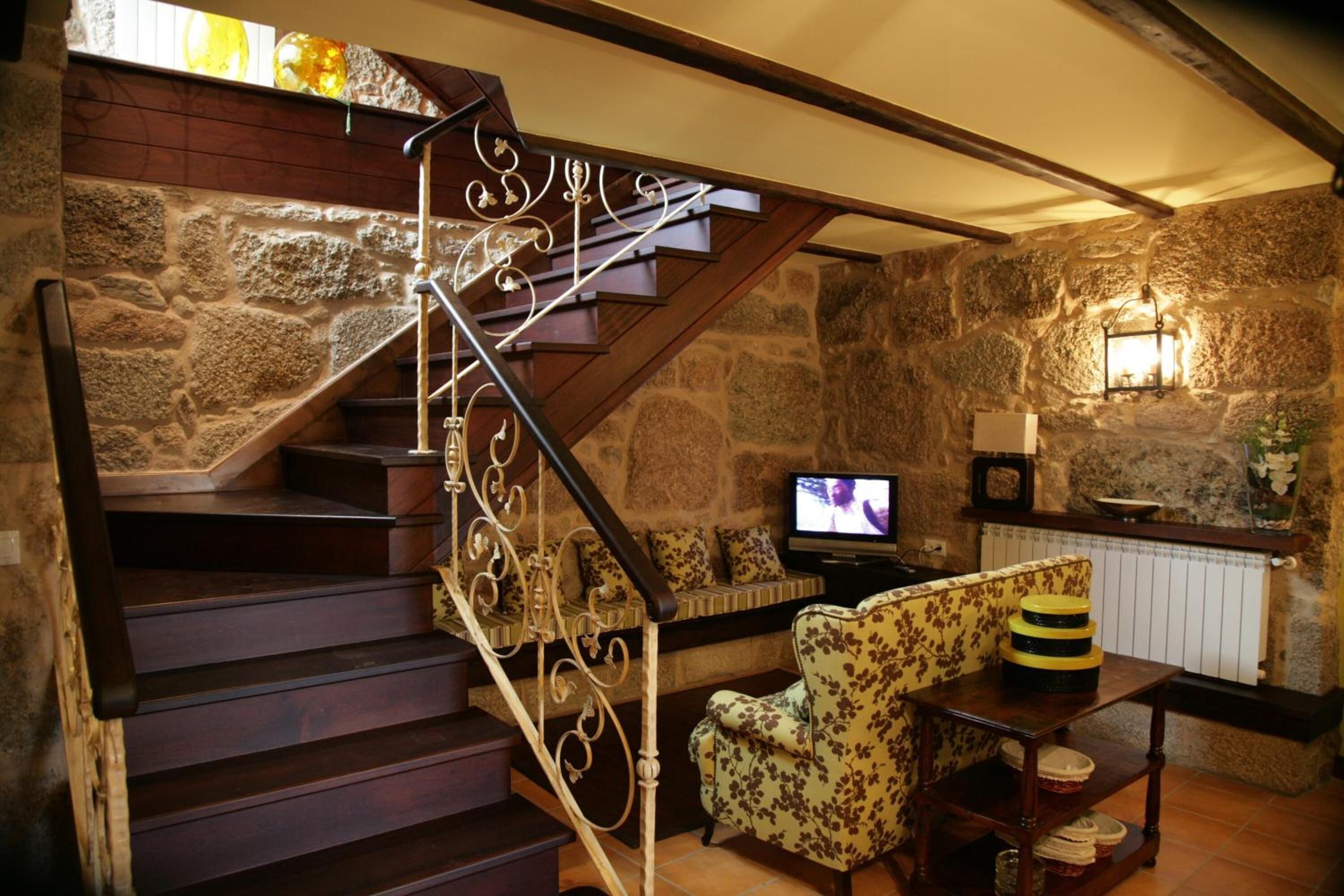 Una habitación con paredes de piedra, una escalera de madera, un televisor, un sillón floral y una mesa con adornos.