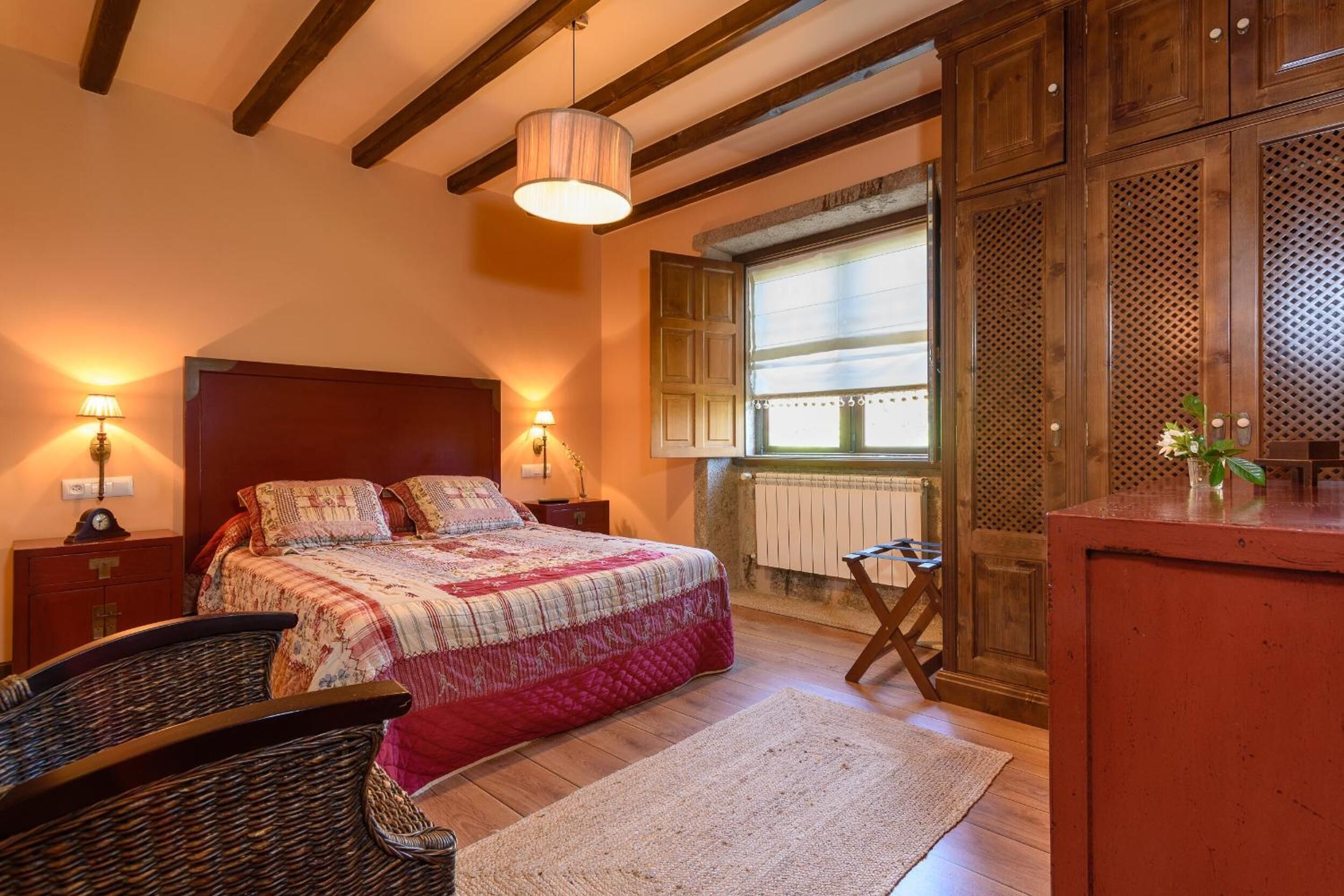 Quarto com cama de madeira, duas luminárias de cabeceira, janela com persianas, radiador e uma cadeira de madeira.