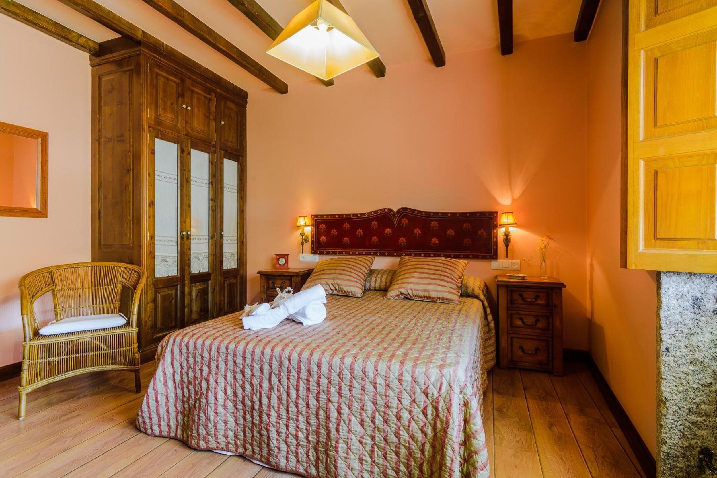 A bedroom with a large bed, wooden wardrobe, wicker chair, and bedside tables with lamps.
