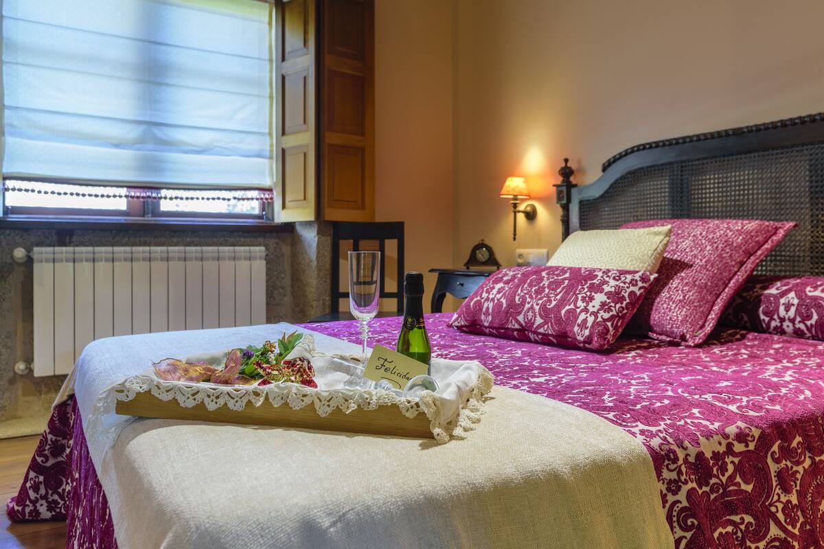A neatly made bed with a tray of food and a bottle of champagne, a bedside lamp, and a window with blinds.