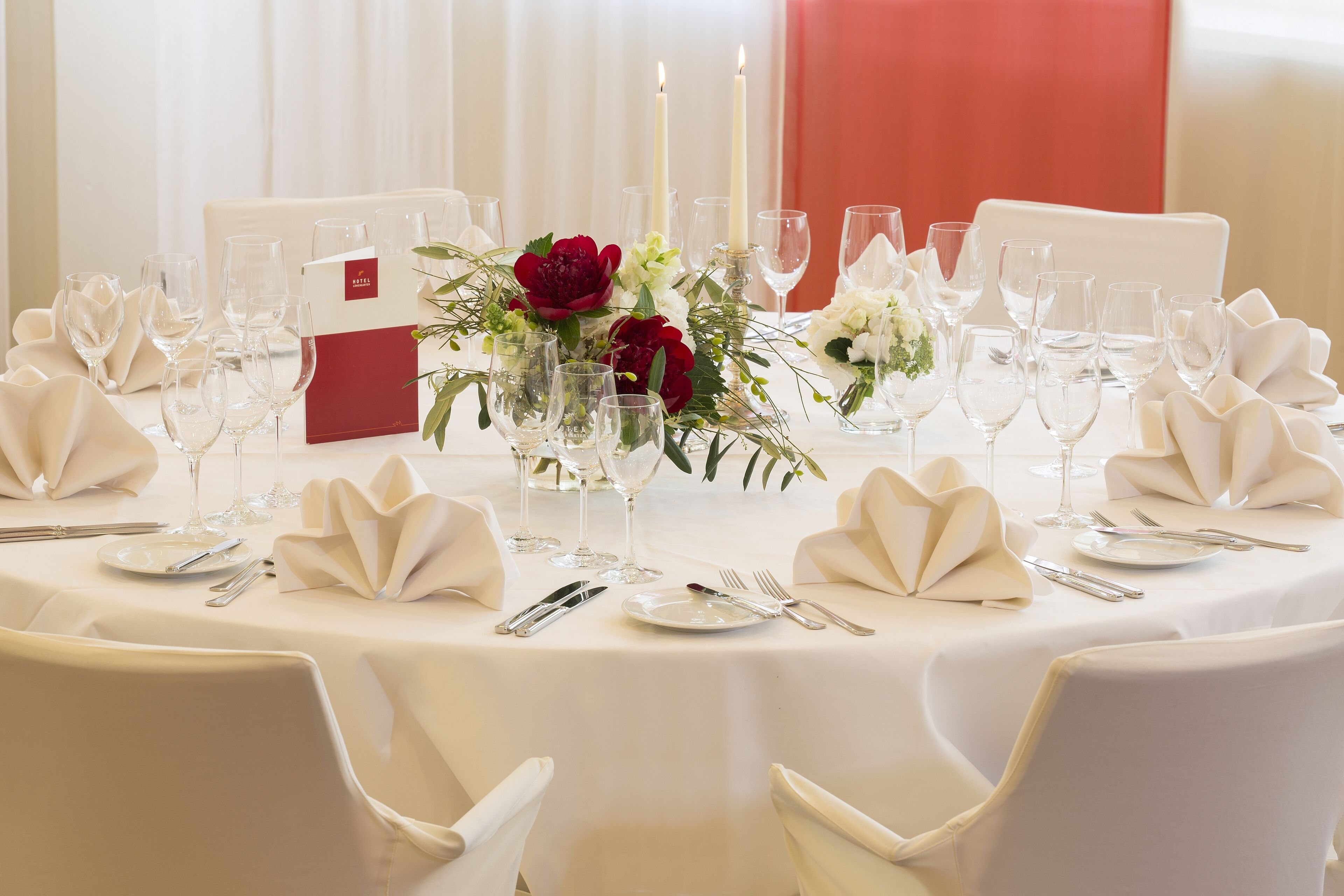 A round table set for a formal event with white tablecloths, folded napkins, and a centerpiece of red flowers.