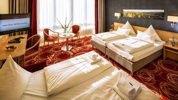 A hotel room with two beds, a desk, a chair, a TV displaying a welcome message, and a painting on the wall.