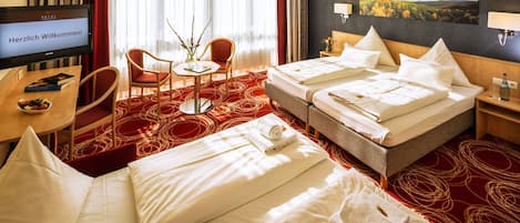 Quarto de hotel com duas camas, uma escrivaninha, uma cadeira, uma TV exibindo uma mensagem de boas-vindas e um quadro na parede.