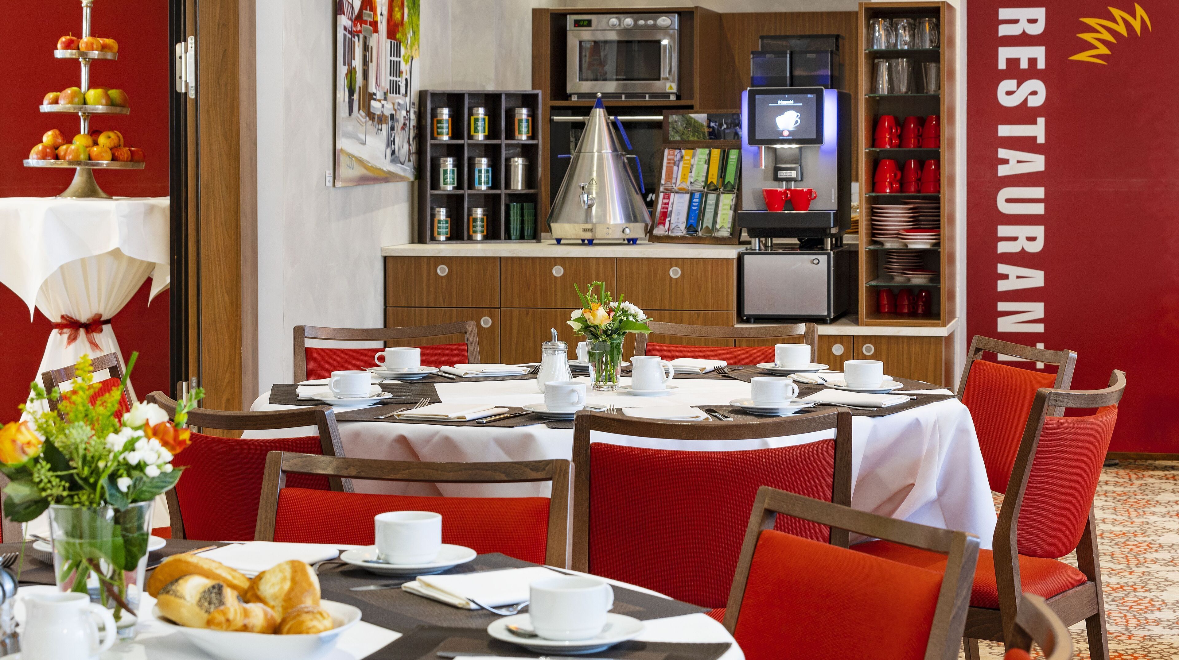Un ristorante con tavoli rotondi, sedie rosse e tovaglie bianche. Sui tavoli si vedono diversi piatti, tra cui pasticcini e un'esposizione di frutta.