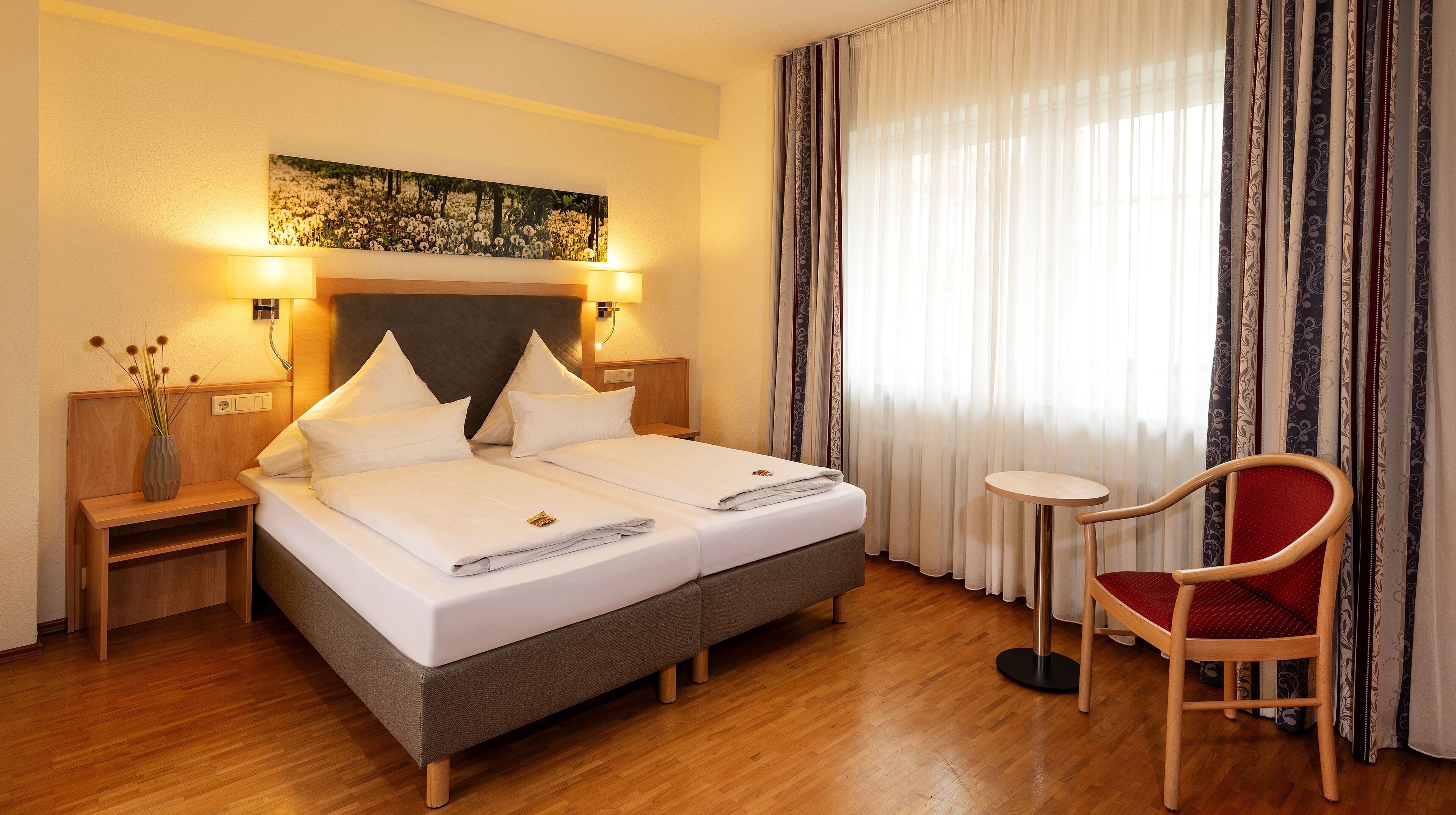 Ein Hotelzimmer mit einem Doppelbett, zwei Nachttischen, einem kleinen runden Tisch, einem roten Sessel und einem großen Fenster mit Vorhängen.