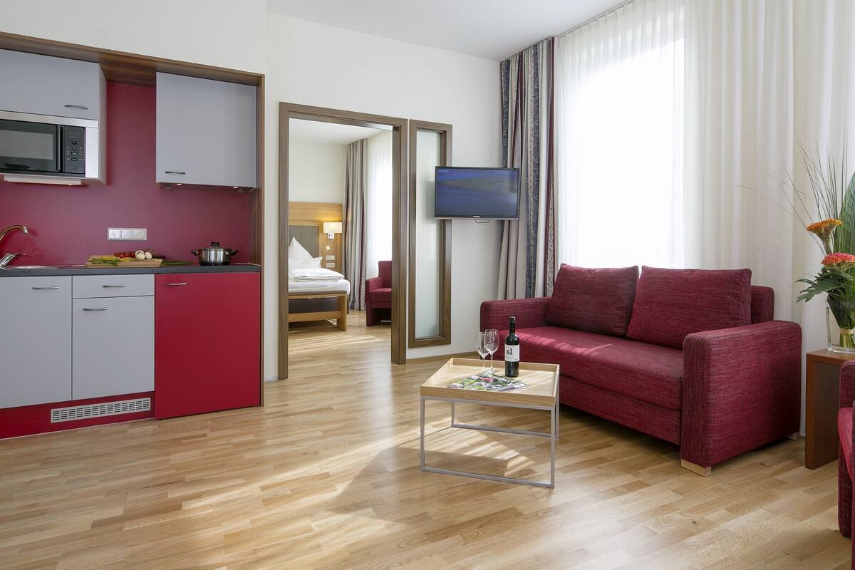 A compact hotel room with a kitchenette, a red sofa, a small coffee table, and a TV.